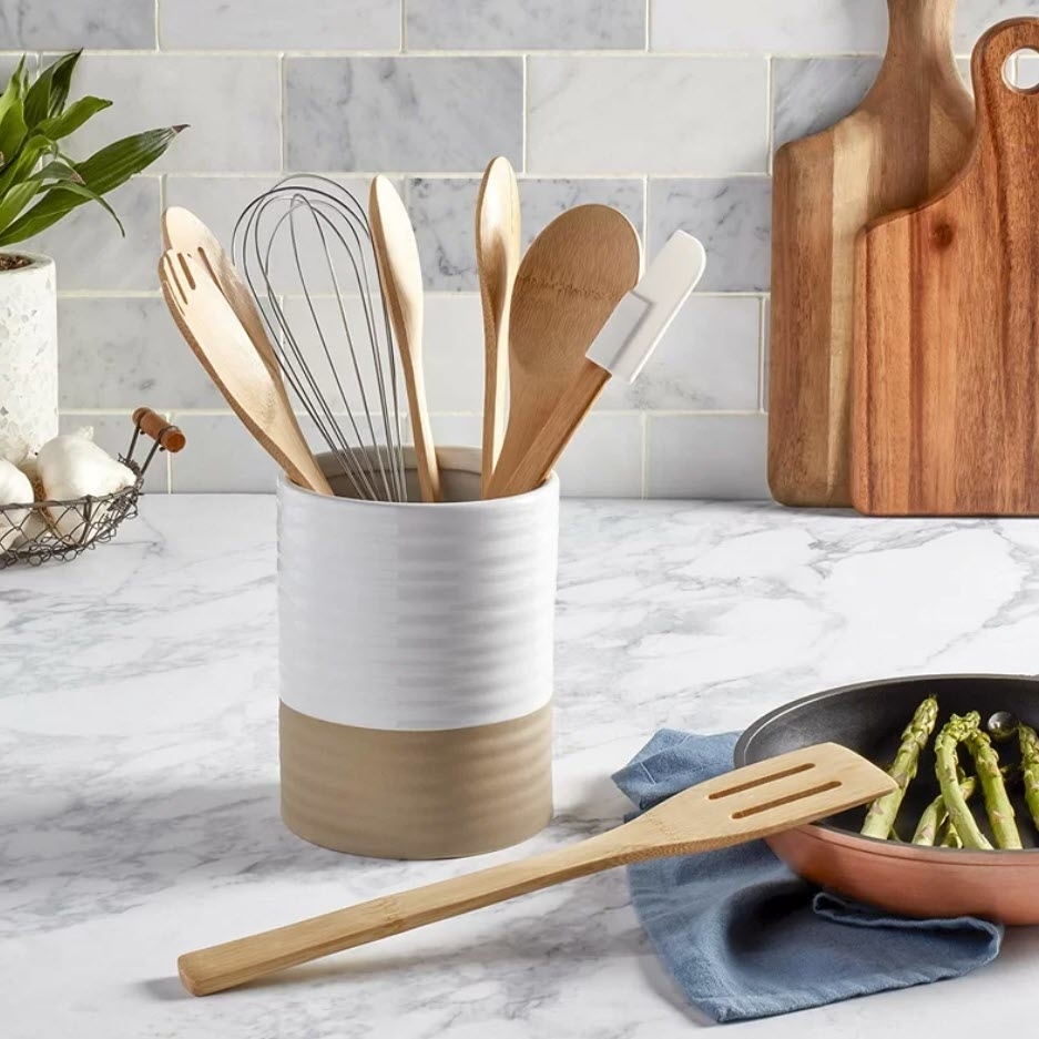 Kitchen utensils including a whisk and wooden spoons in a two-tone holder on a countertop with a cutting board and a pan with asparagus behind
