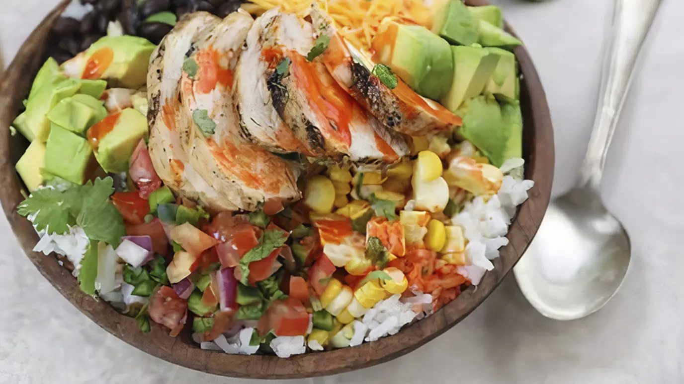 Sliced chicken and avocado atop a bed of rice with mixed vegetables in a bowl