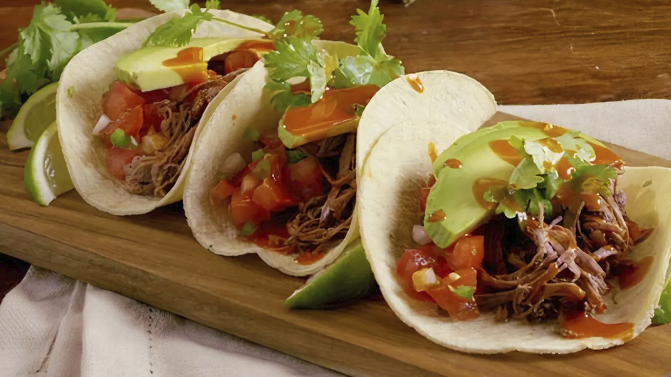 Three tacos with shredded meat, avocado slices, and diced tomato on a wooden tray
