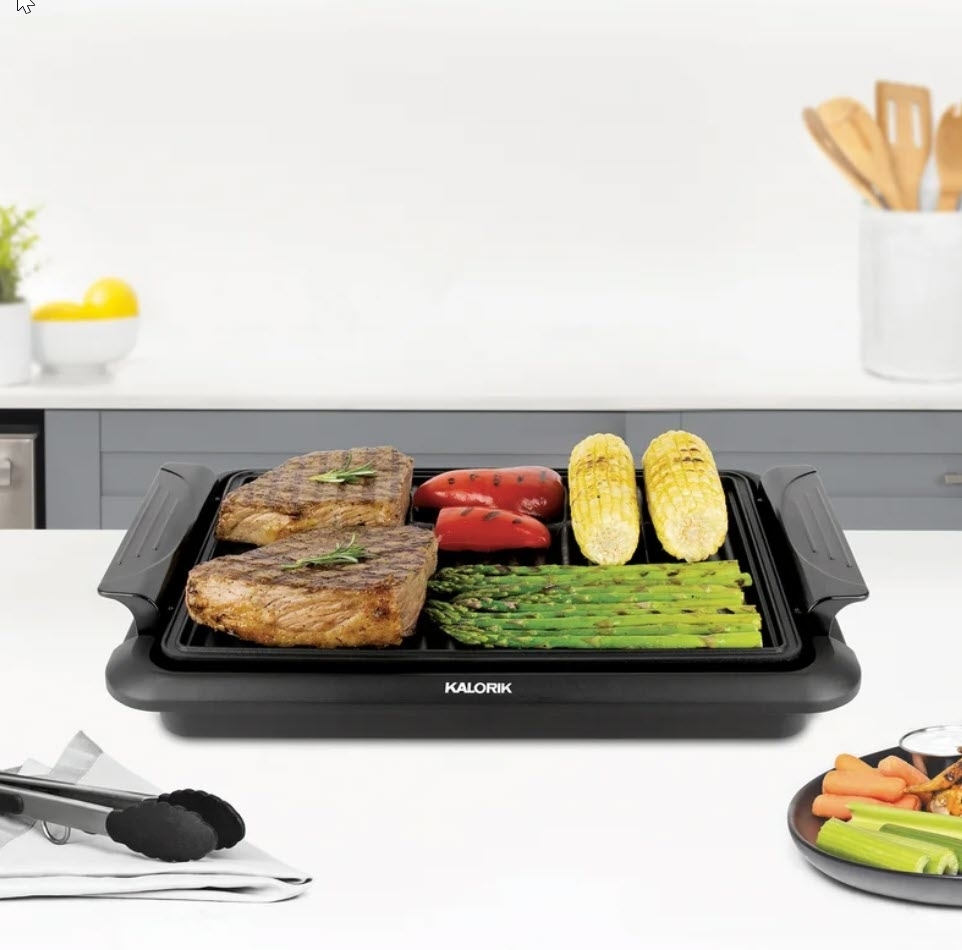 Indoor electric grill on kitchen counter cooking steaks, vegetables, and corn