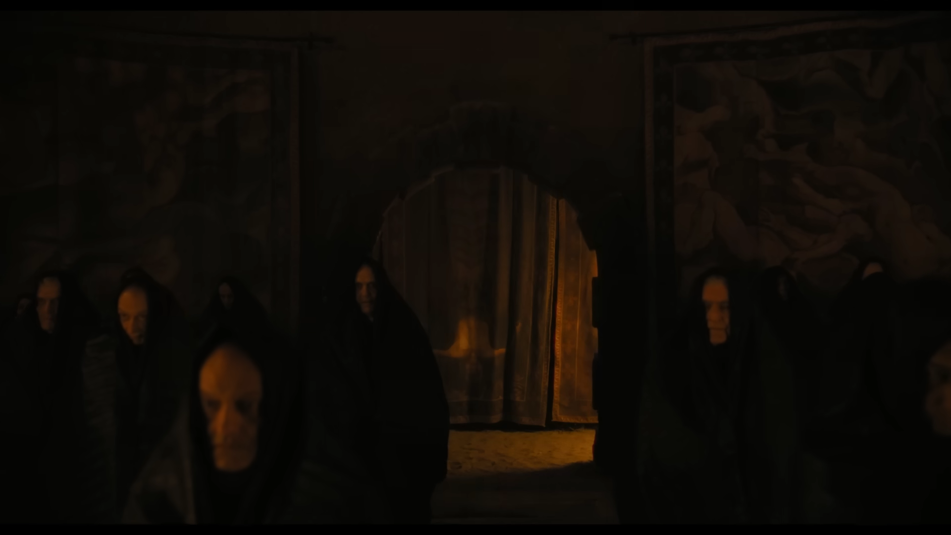 Dark room with people in cloaks standing solemnly by stone walls, tapestries in the background