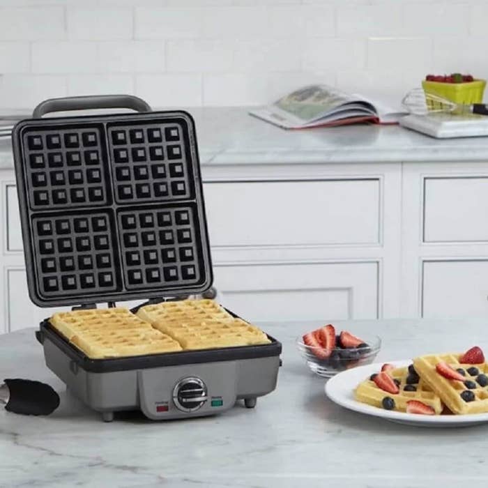 Waffle iron open on a counter with fresh waffles and a plate of waffles topped with fruit nearby