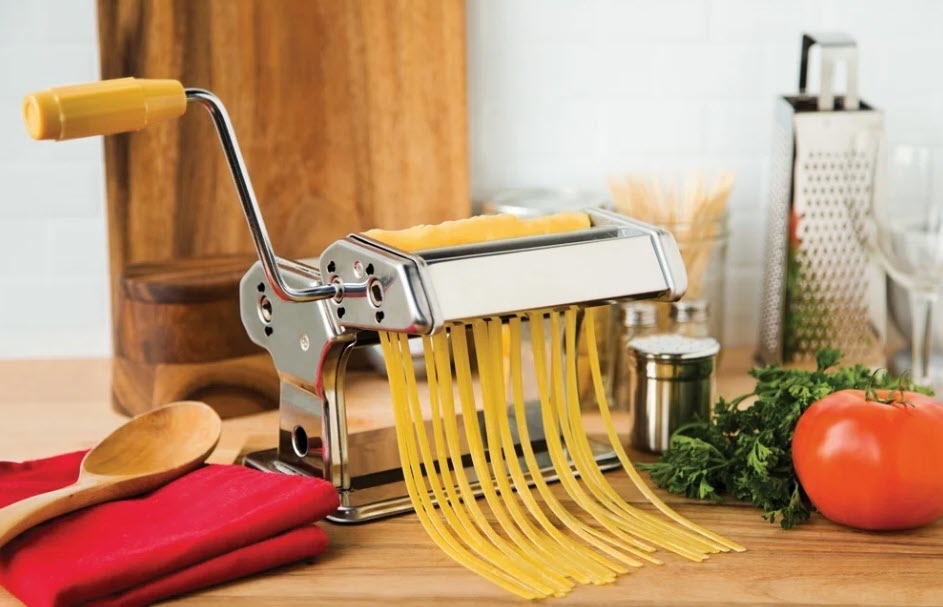 Pasta maker clamped to counter extruding fresh spaghetti noodles next to a tomato and utensils