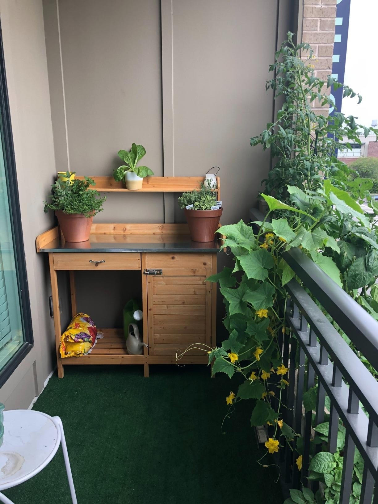 Wooden balcony plant station with herbs and blooming plants, artificial grass flooring, and gardening supplies