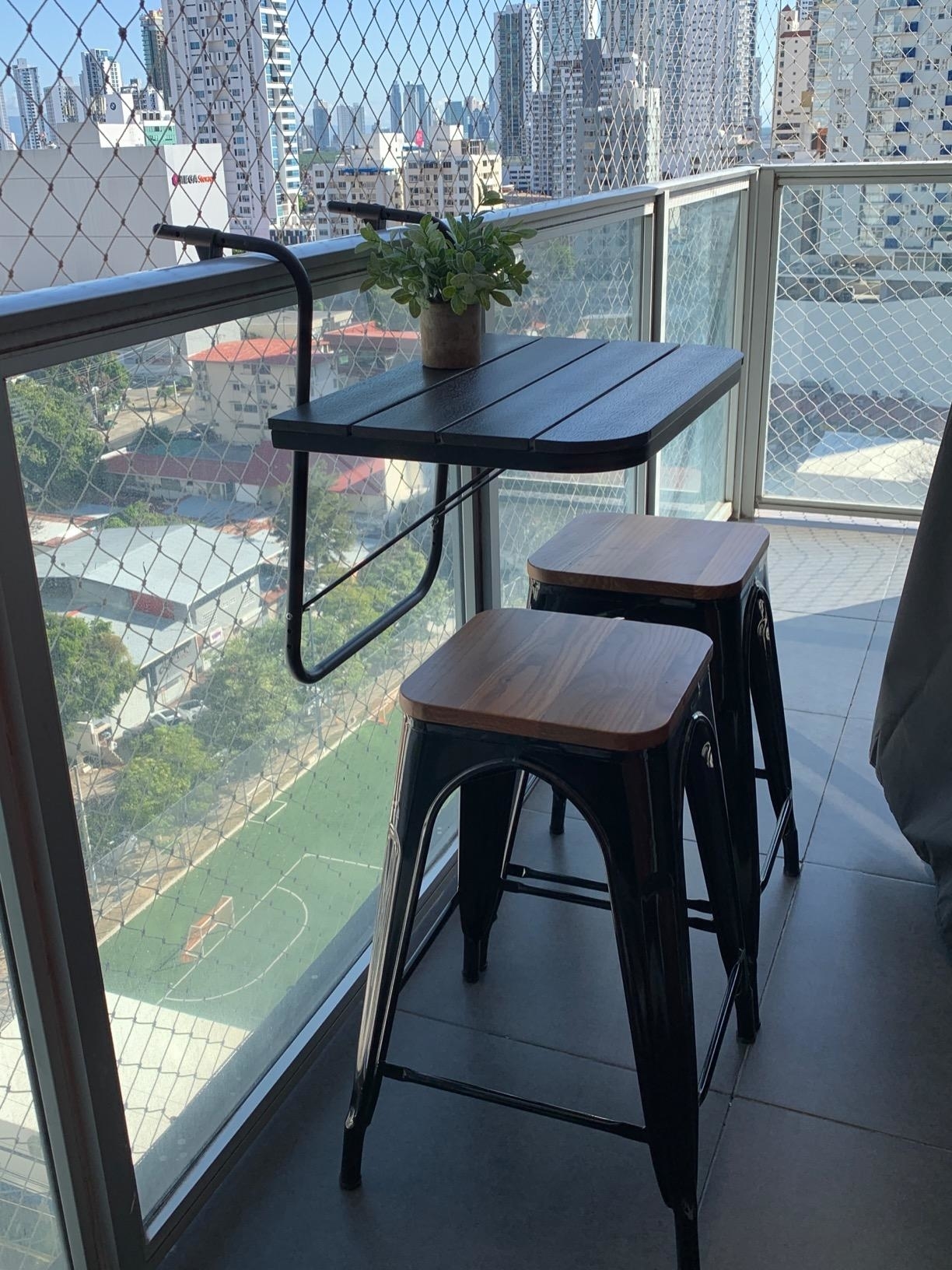 Small balcony with a black folding table, two wooden stools, and a potted plant, overlooking a cityscape