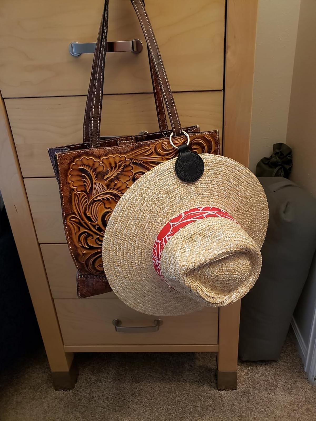 A straw hat with a red band hangs on a cabinet knob beside a tooled leather tote bag