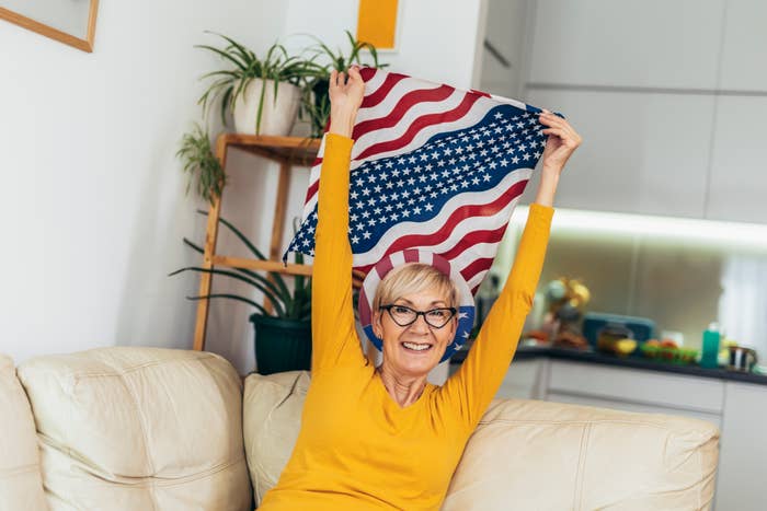 Elderly woman smiling, holding an American flag indoors, sitting on a beige couch with houseplants in the background