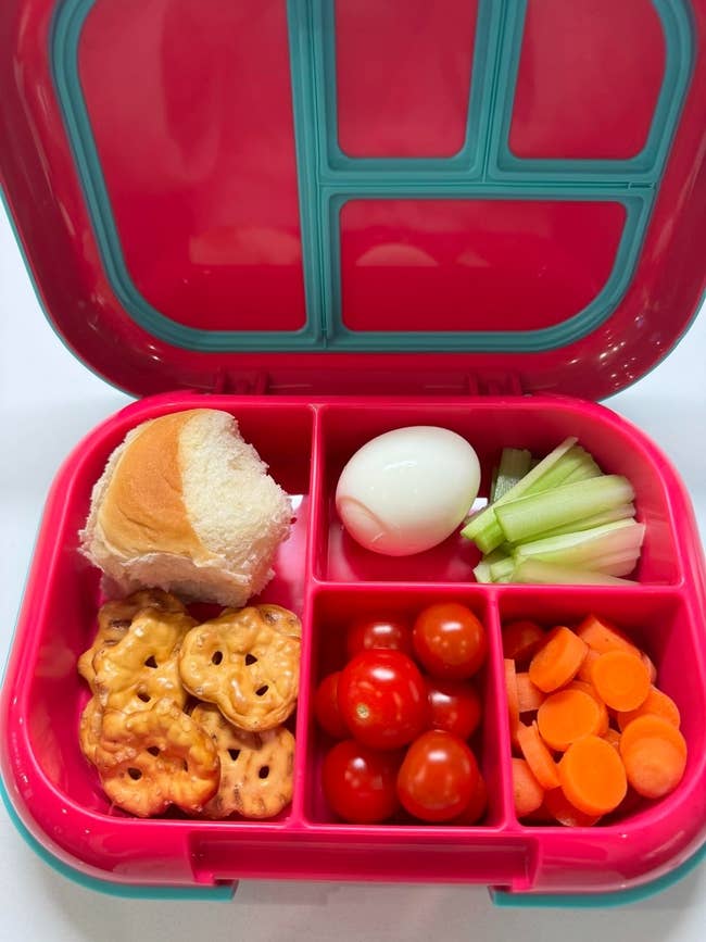 The bento lunchbox unfastened  with a roll, pretzels, boiled egg, tomatoes, carrots, and celery sticks.
