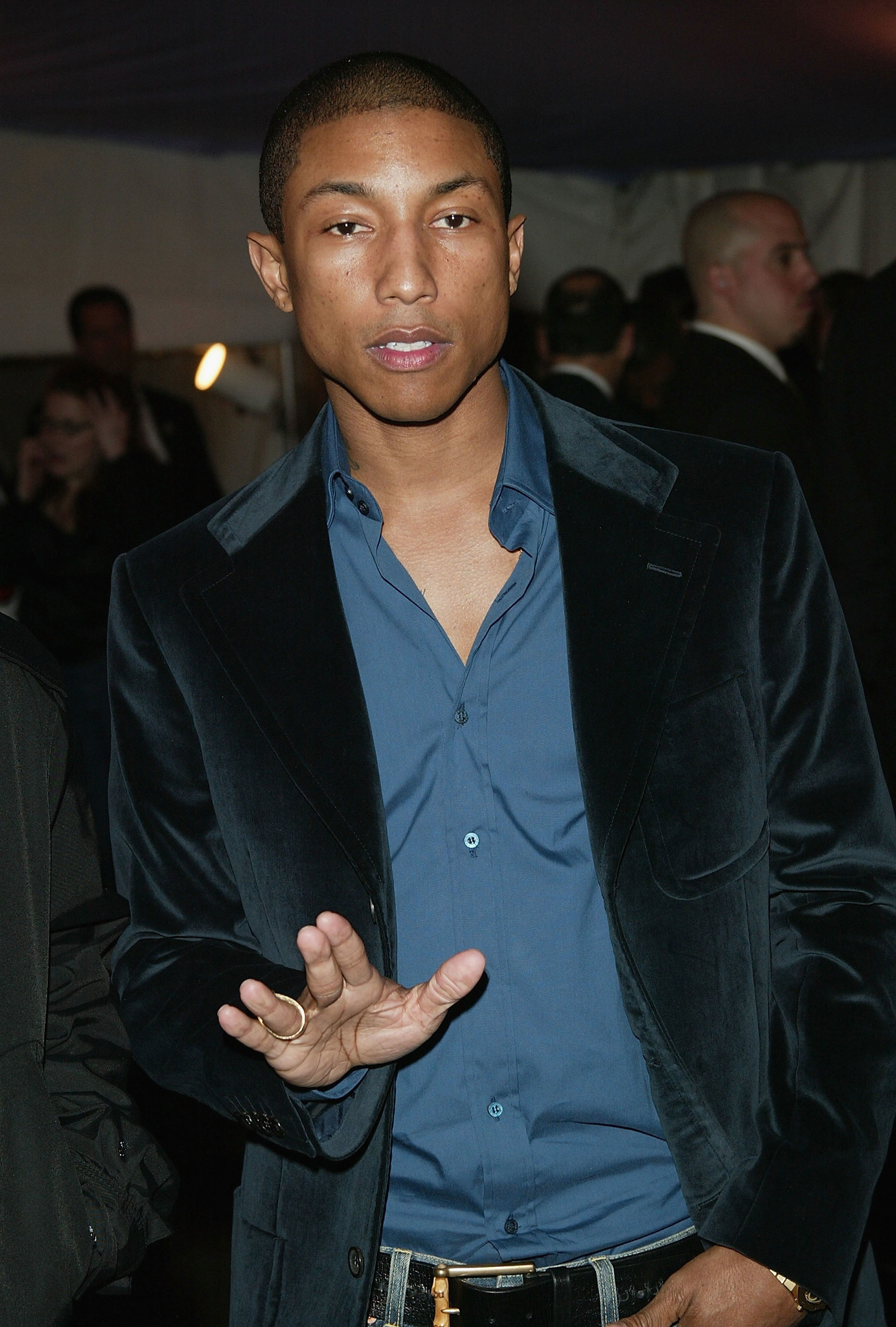 Pharrell Williams in a velvet jacket and collared shirt, gesturing with his hand, at an event