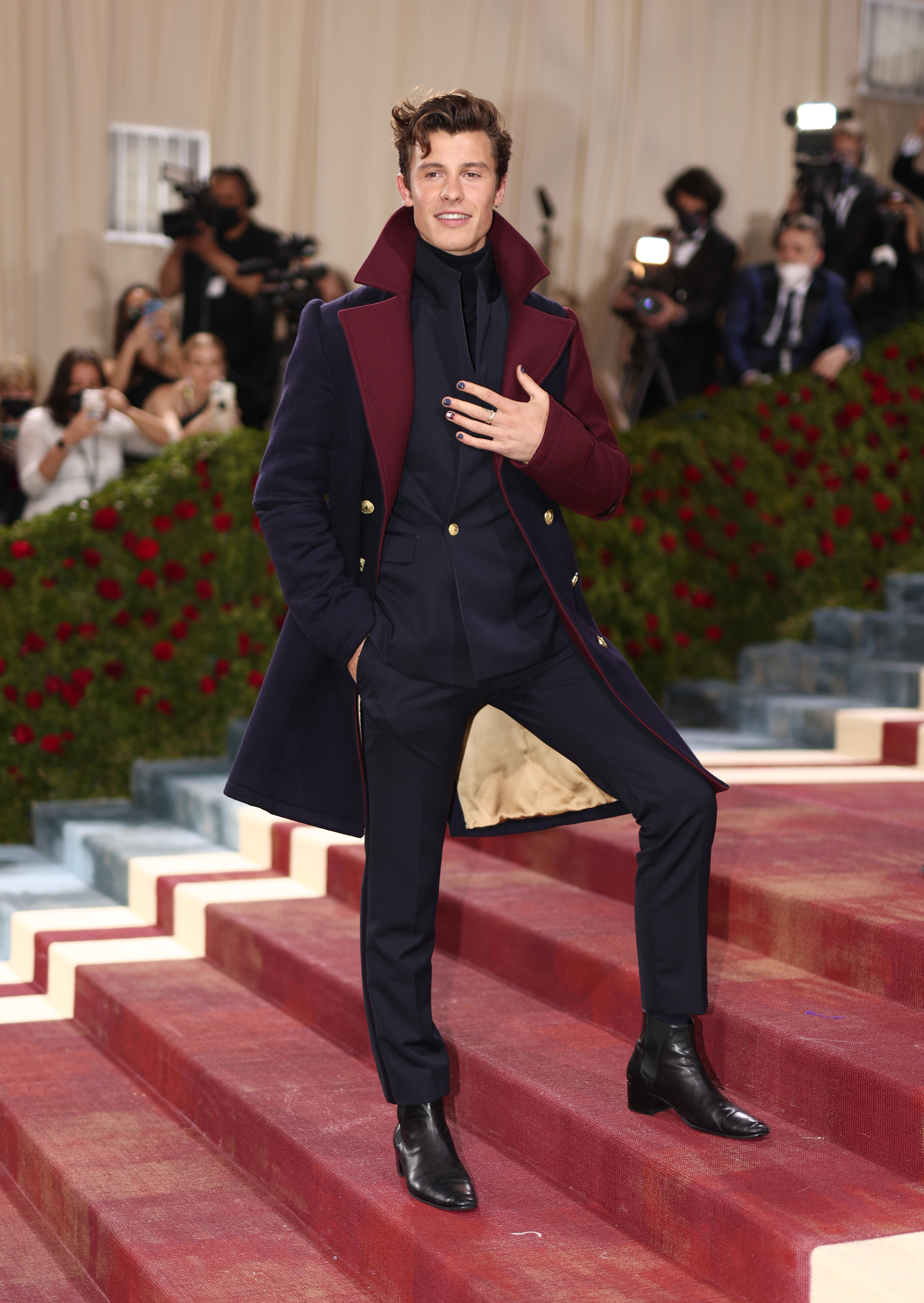 Man on red carpet posing in a layered navy outfit with a coat, scarf, and black boots