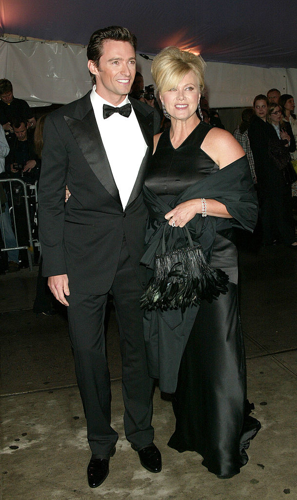 Two individuals posing, one in a black tuxedo and the other in a black off-shoulder gown with feather details