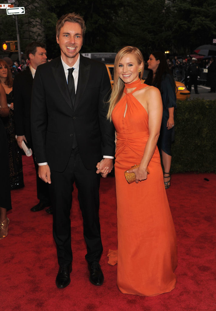 Two celebrities smiling and holding hands on the red carpet, with the man in a black suit and the woman in an orange sleeveless gown