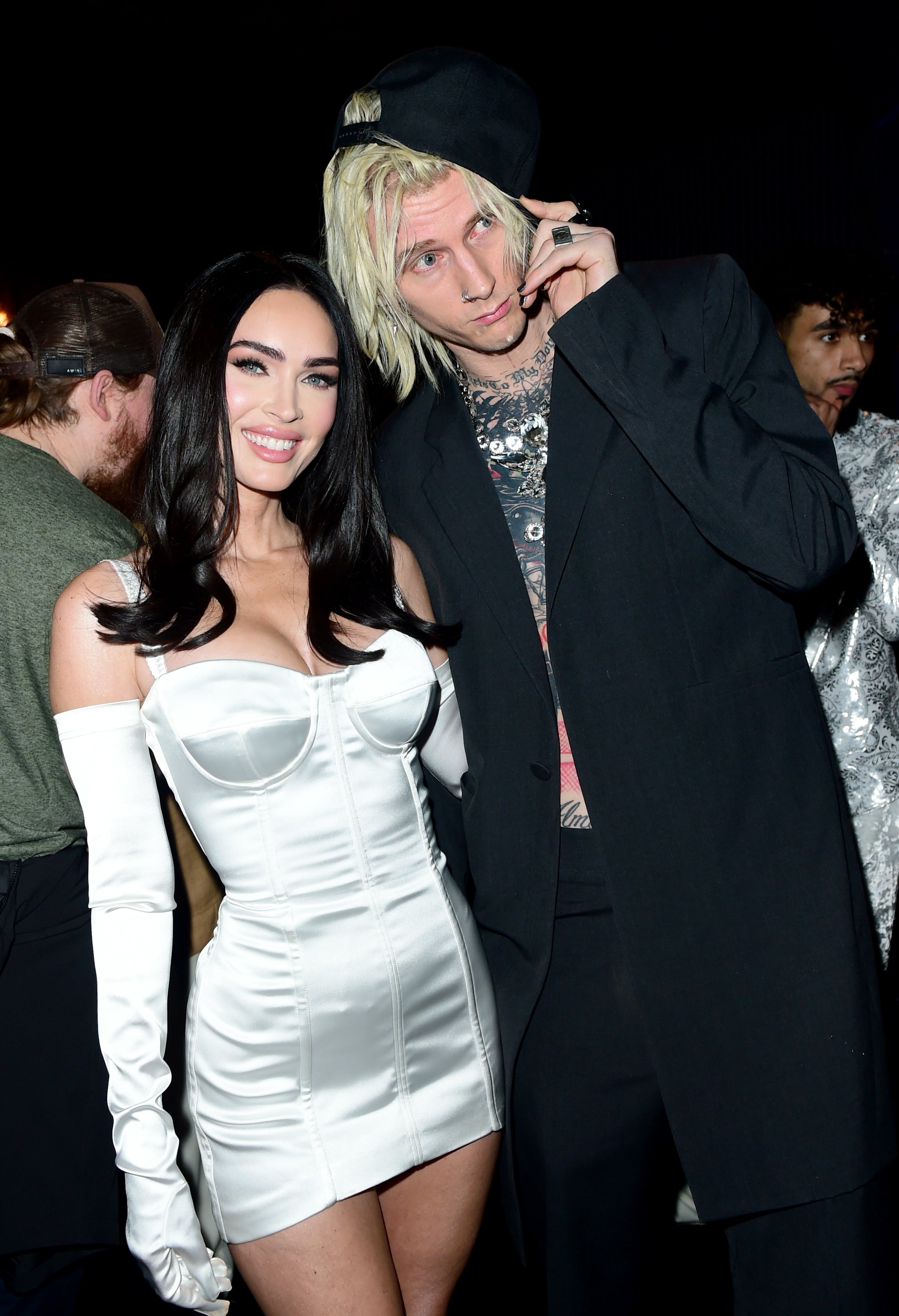 Megan Fox in a corset dress with gloves, and Machine Gun Kelly in a suit with silver chain necklaces at an event