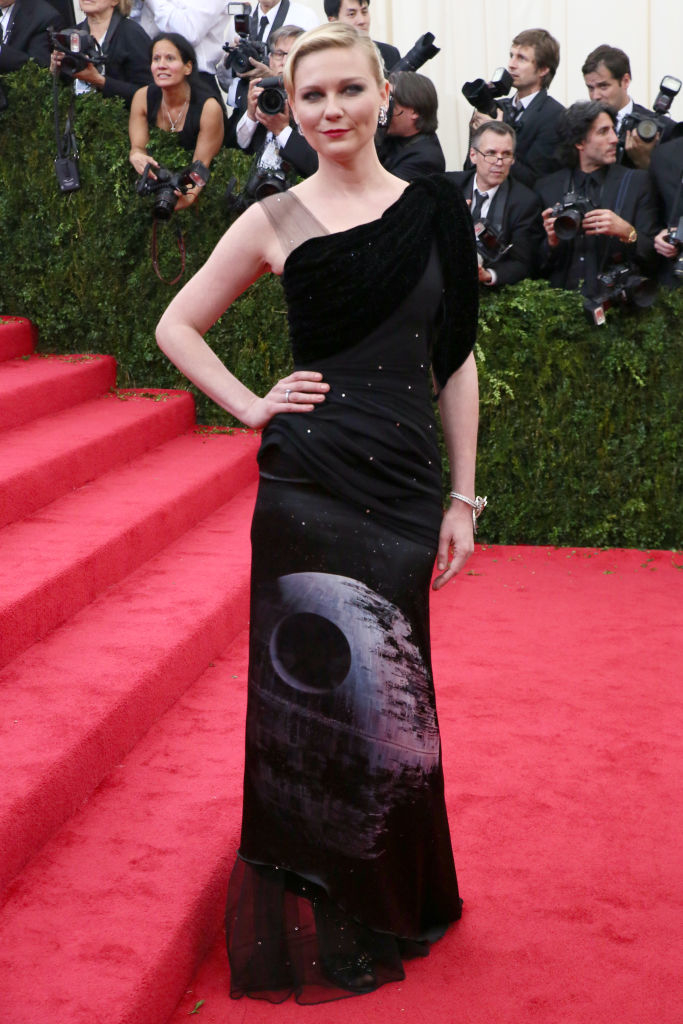 A woman on the red carpet in a black one-shoulder gown with a moon print, standing with hand on hip. Photographers in the background