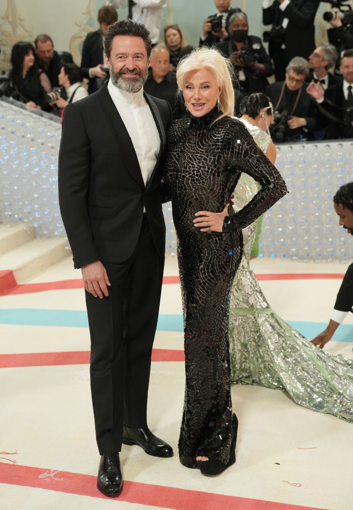 Two people posing, one in a black suit and the other in a fitted, shimmering gown with a train