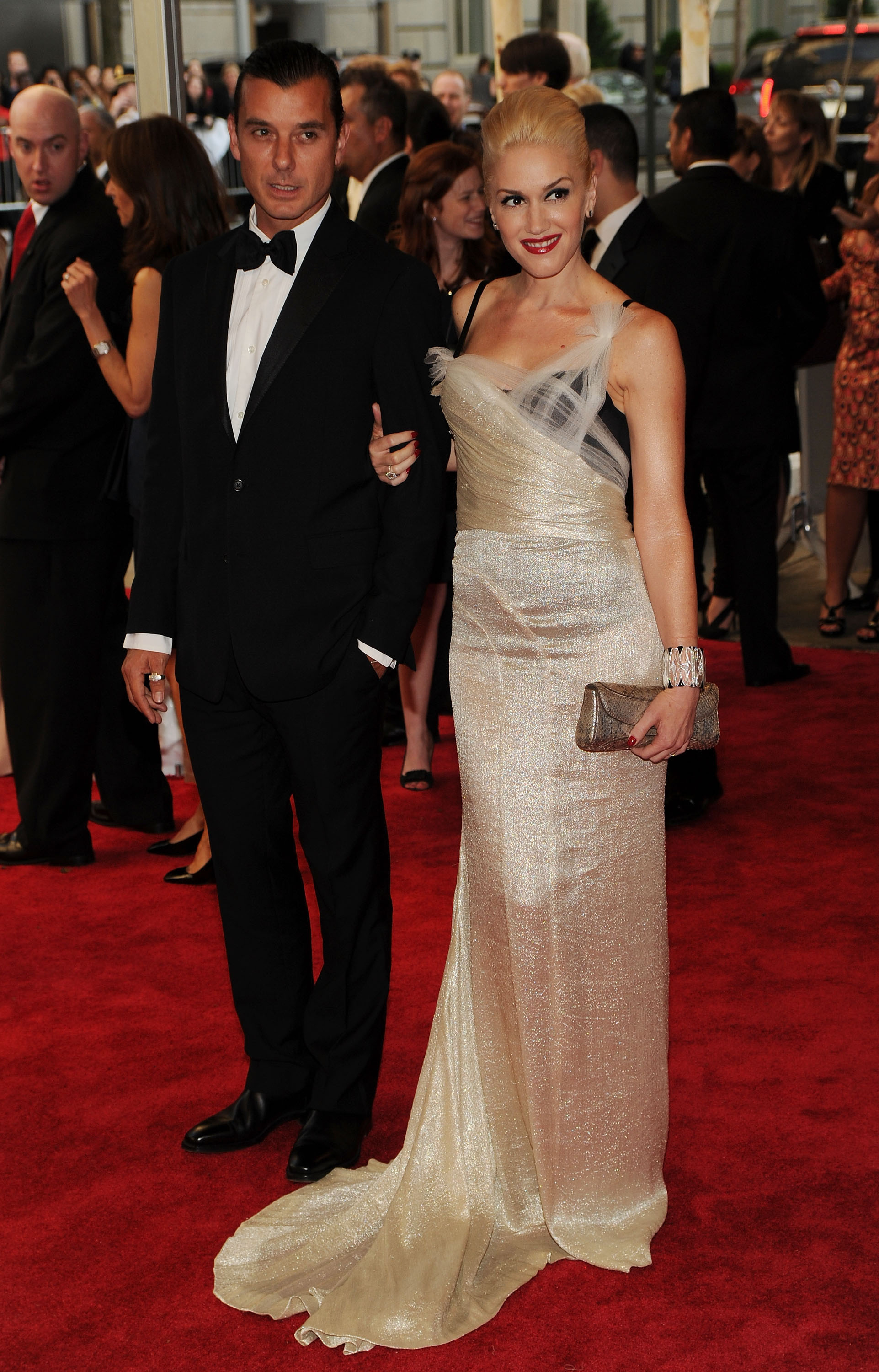Two celebrities on red carpet; man in a suit and woman in a one-shoulder gown with a clutch