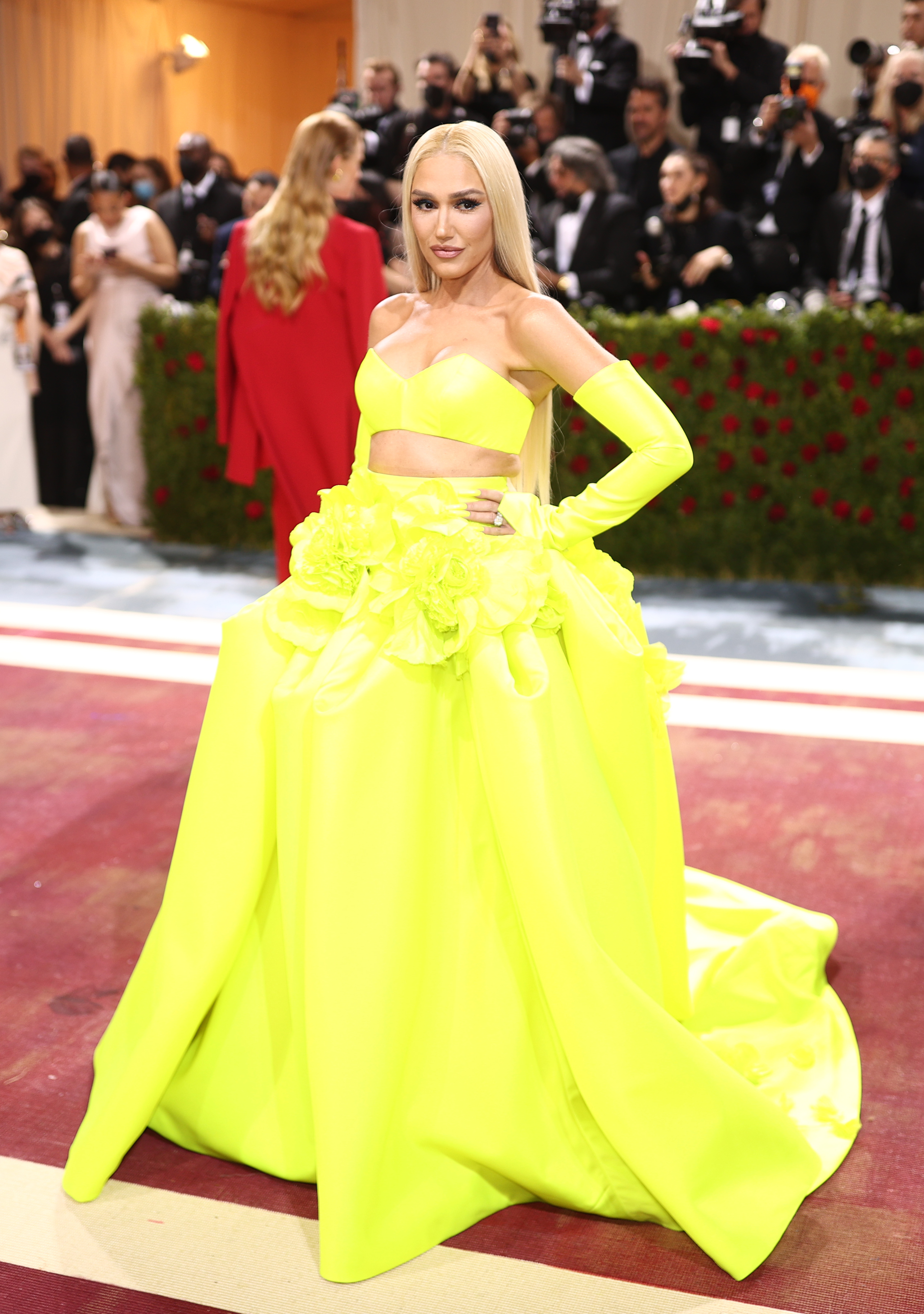 Woman in striking yellow gown with midriff cutout, long sleeves, and floral accents posing on event carpet
