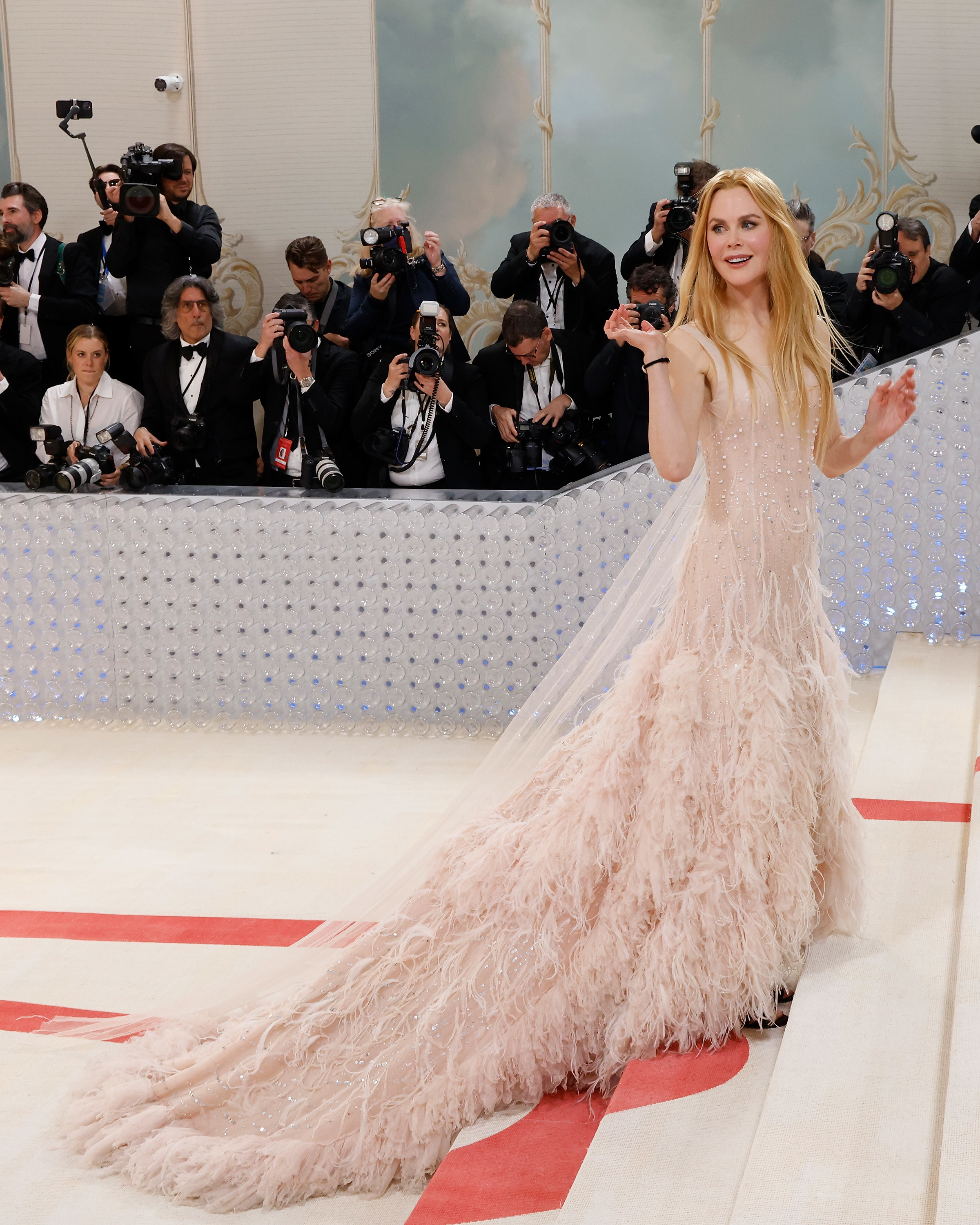 Nicole Kidman in an elegant gown with a long feathery train, waving, with photographers in the background