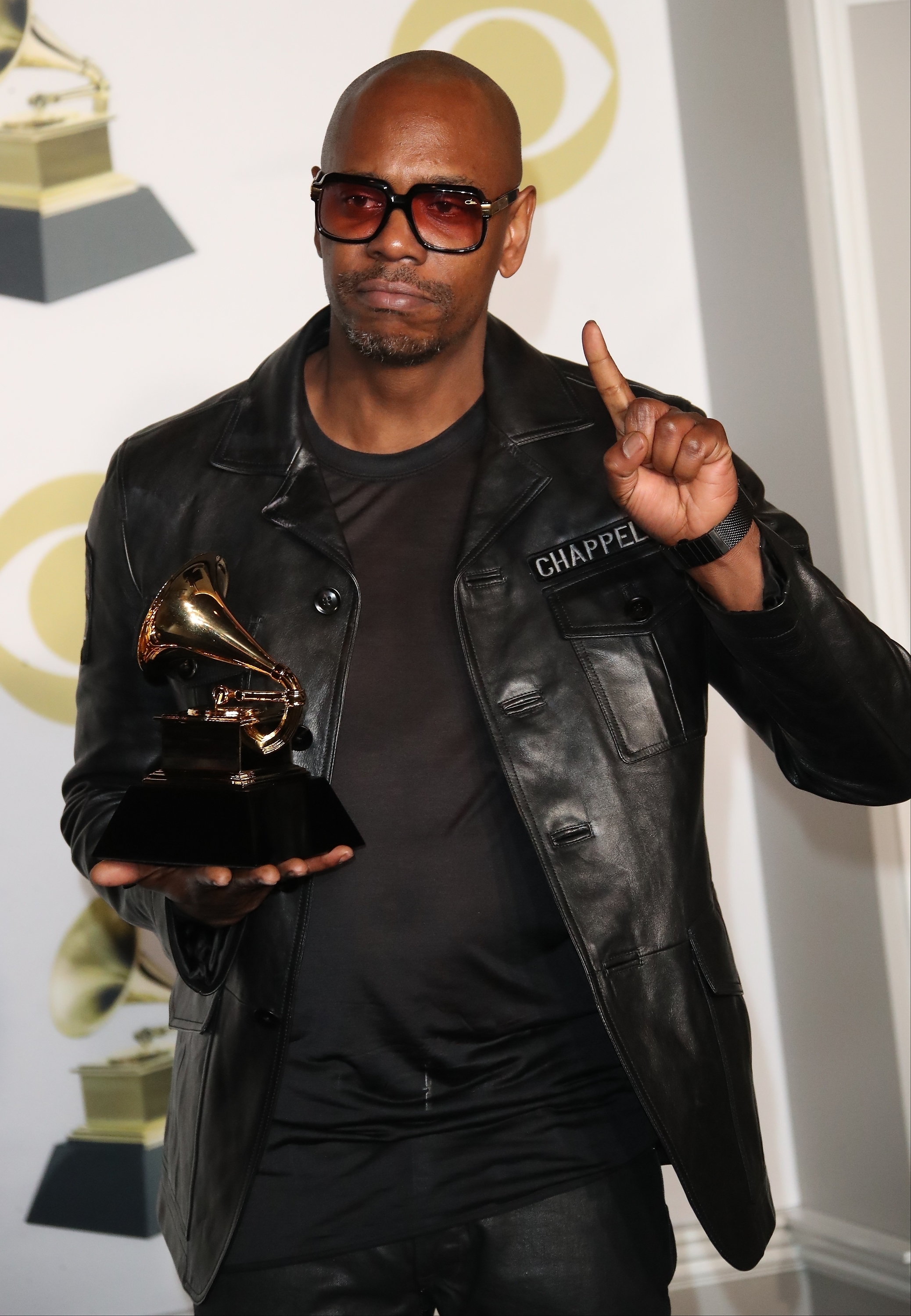 Dave Chappelle in a black leather jacket, holding a Grammy award and posing with one finger raised