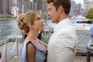 Sydney Sweeney and Glen Powell are close together, with a waterfront and boats in the background. Sydney Sweeney wears a blue formal dress, and Glen wears a suit