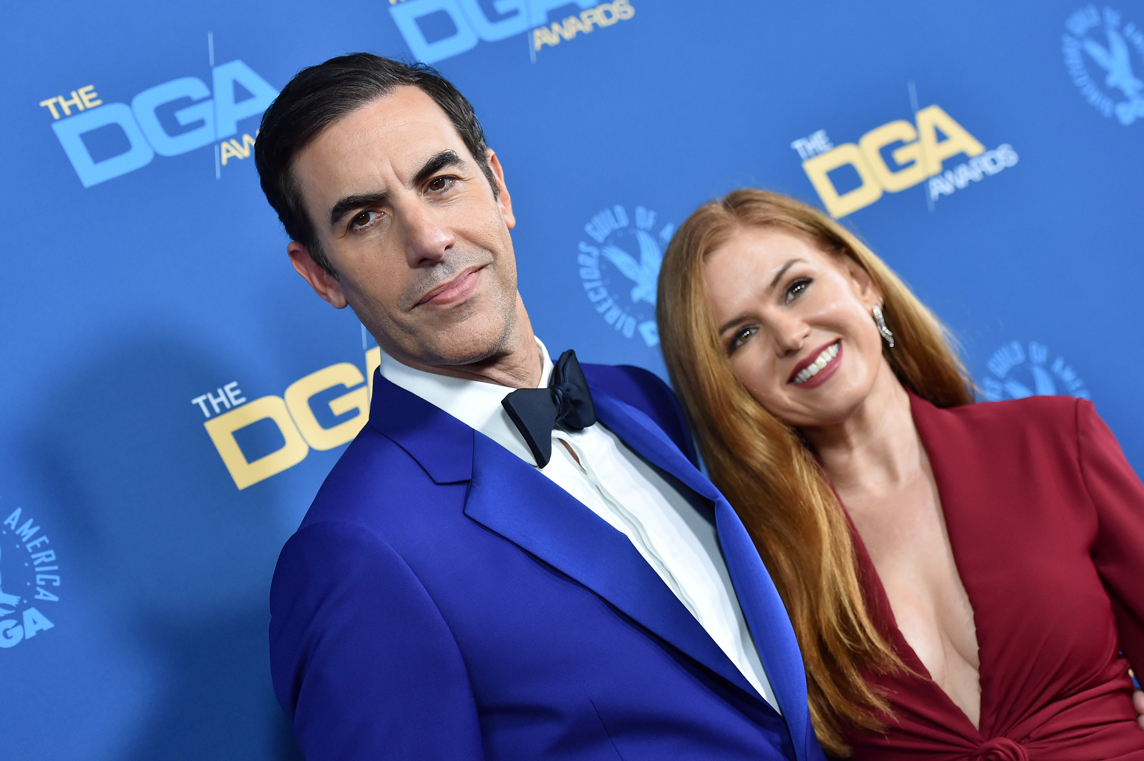 Two celebrities posing; one in a blue suit and bow tie, the other in a red dress, at the DGA Awards