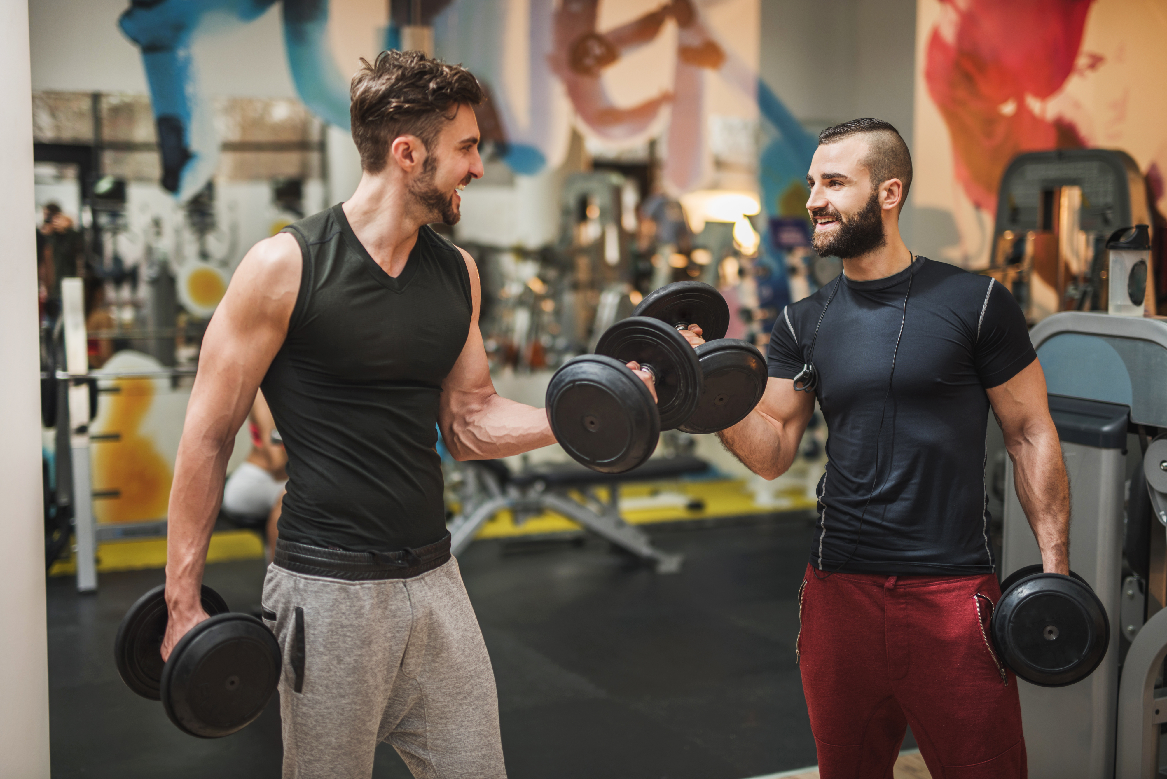 Two men smiling while lifting dumbbells at a gym
