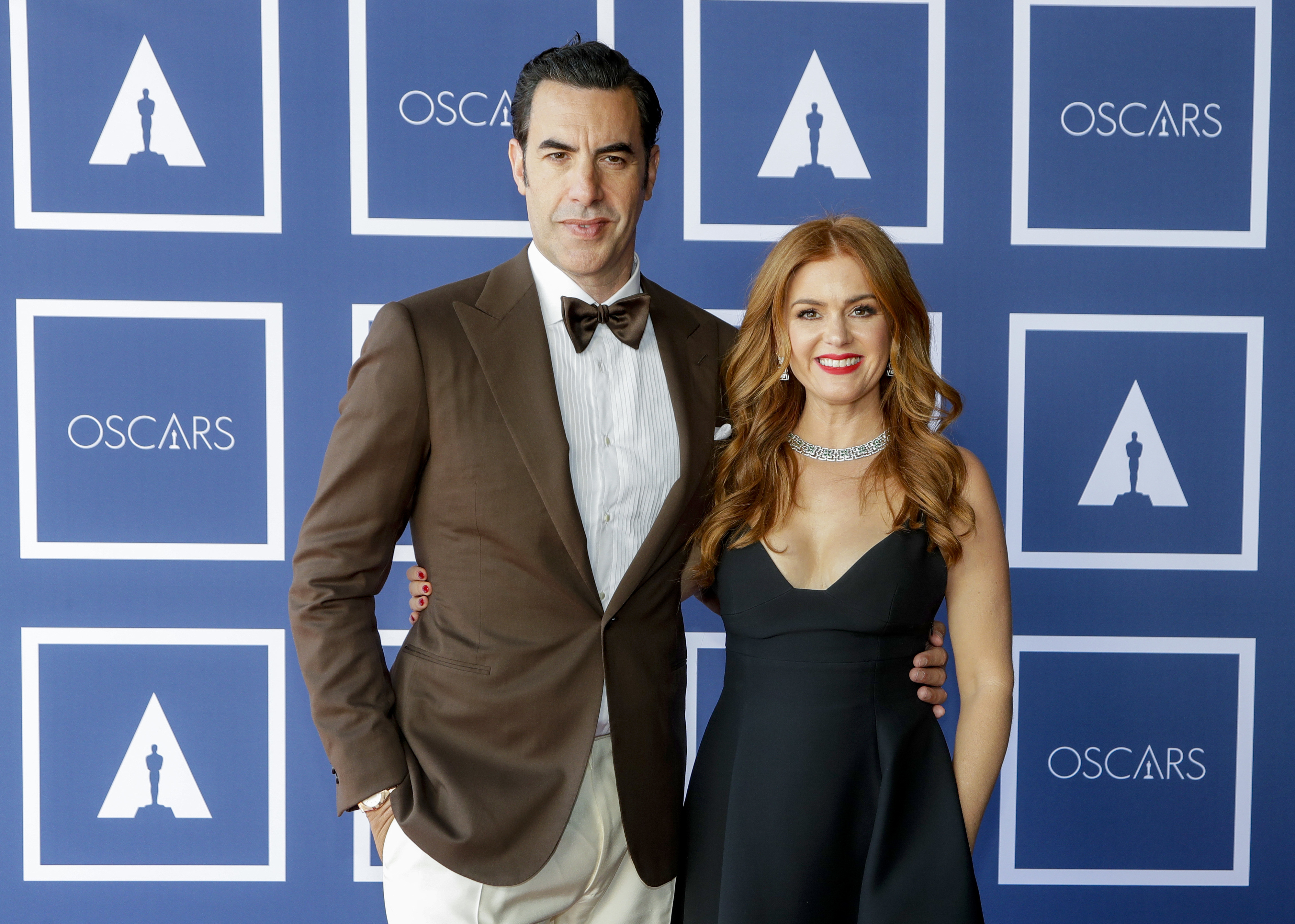Two individuals posing at the Oscars; one in a brown suit and bowtie, the other in a black sleeveless gown and necklace