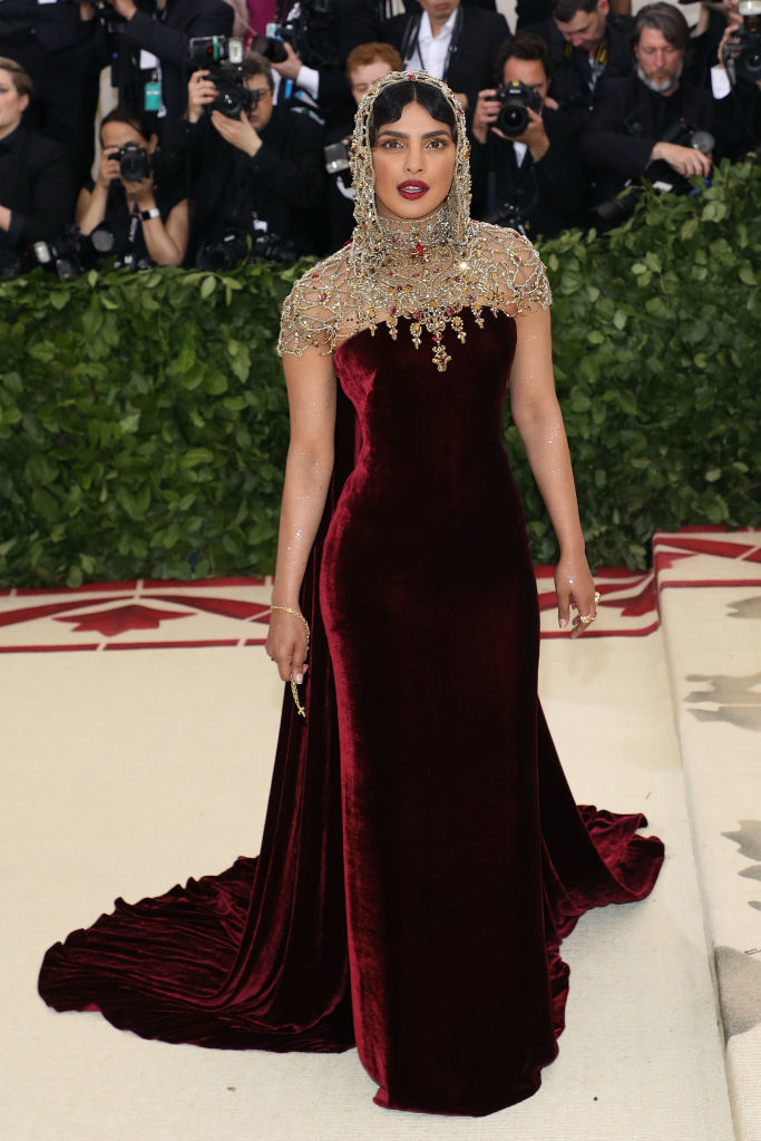 Priyanka standing on a red carpet in a velvet gown with embellished cape and headpiece. Photographers in the background