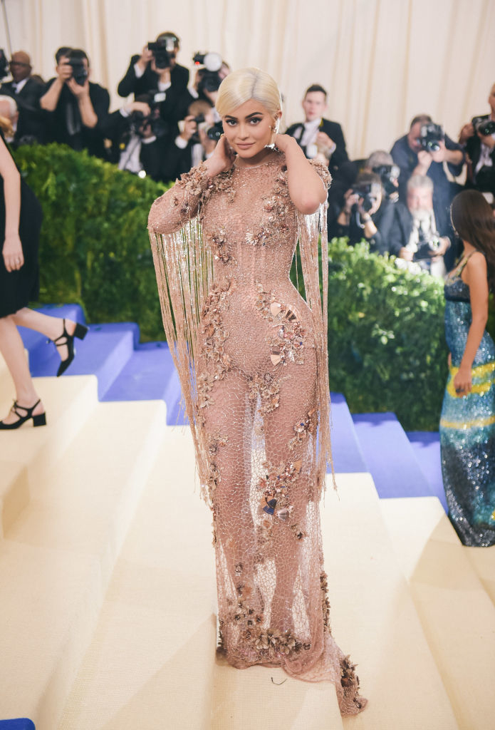 Kylie Jenner in a sheer, embellished gown with fringe details at a gala event. Photographers in the background
