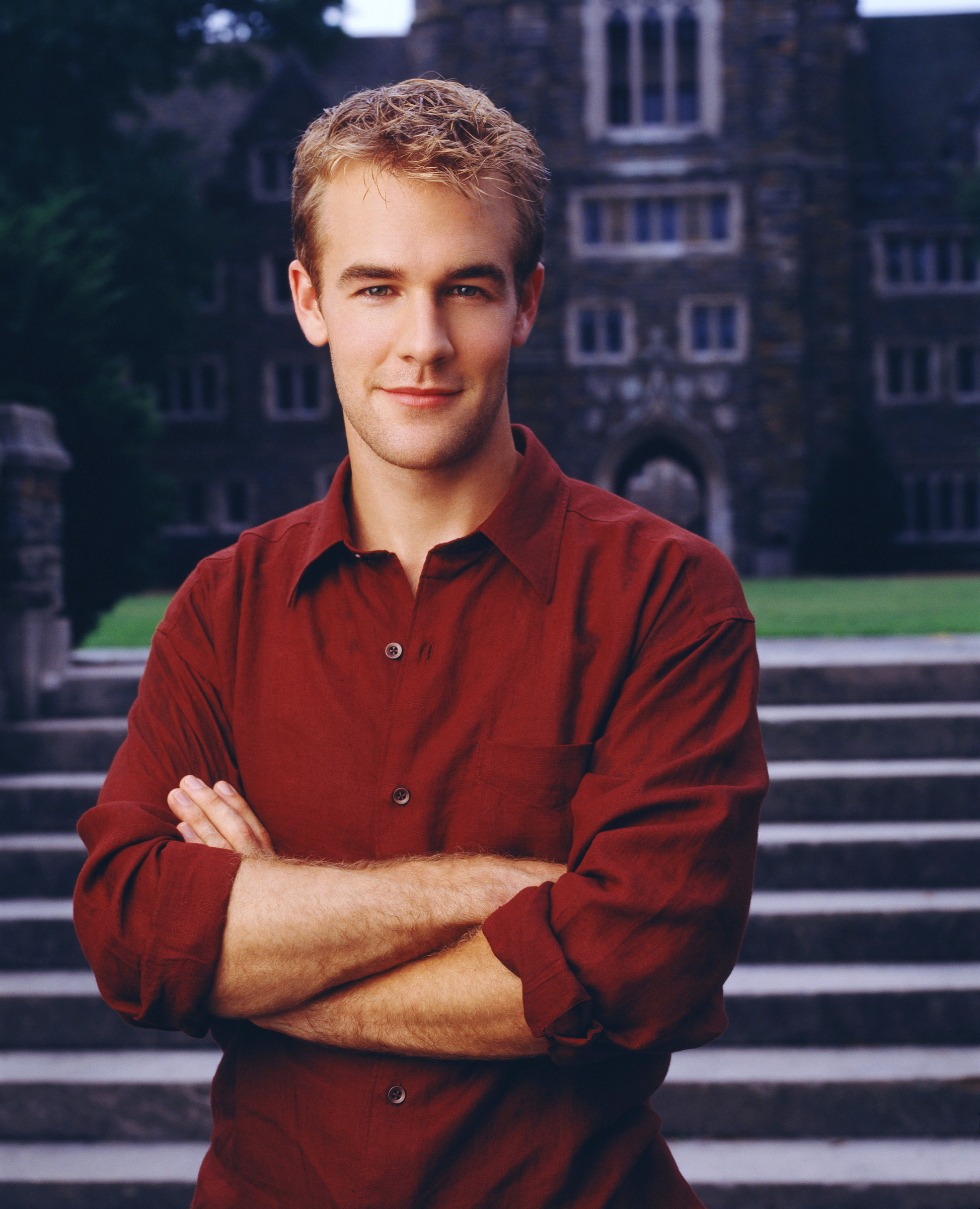James Van Der Beek, as Dawson Leery, stands in front of a building, arms crossed, wearing a casual shirt