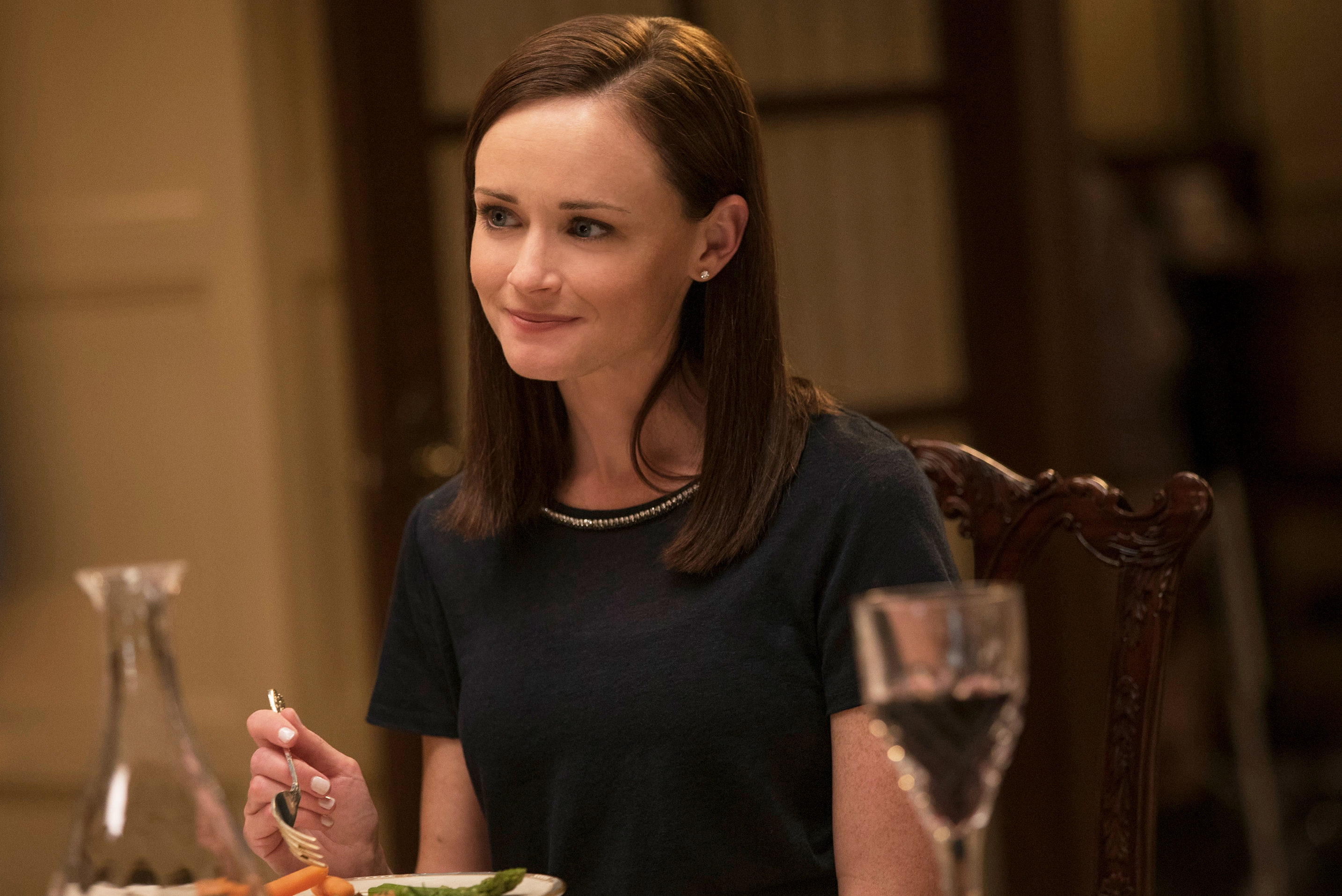 A woman seated at a dining table with a subtle smile, in a casual setting for a TV show scene