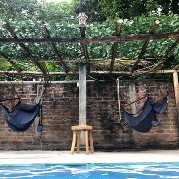 Two empty hammocks beside a pool with a rustic wooden stool under a bamboo canopy