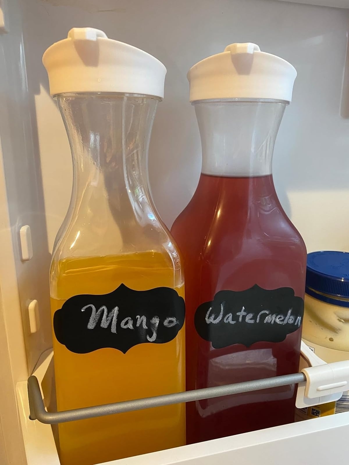 Two labeled carafes with drinks, one marked &#x27;Mango&#x27; and the other &#x27;Watermelon&#x27;, inside a fridge