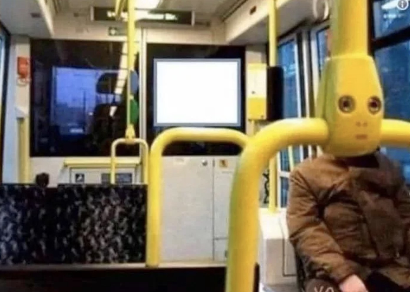 Person in a costume with an elongated head sitting inside a bus next to a screen