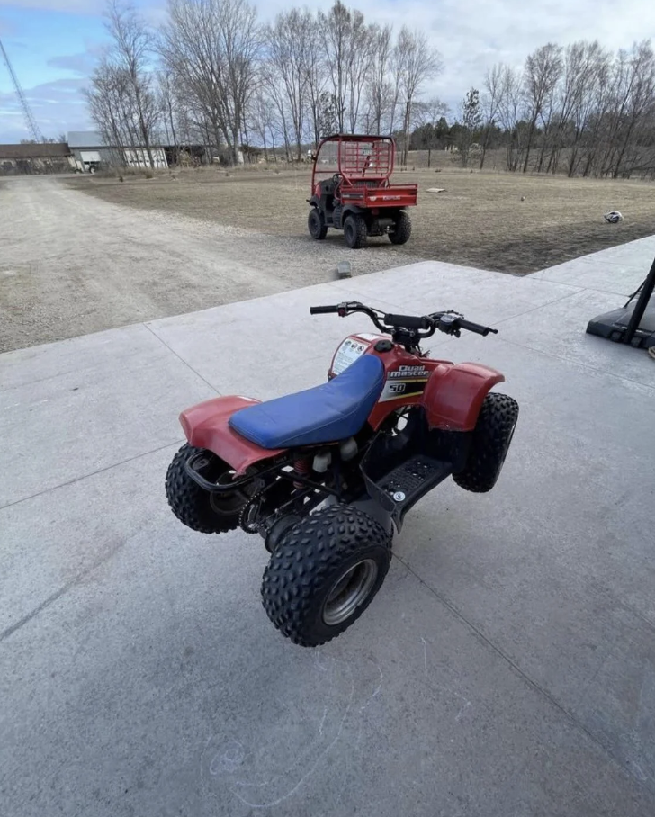 Red and black ATV parked on concrete with an open field and a red utility vehicle in the background