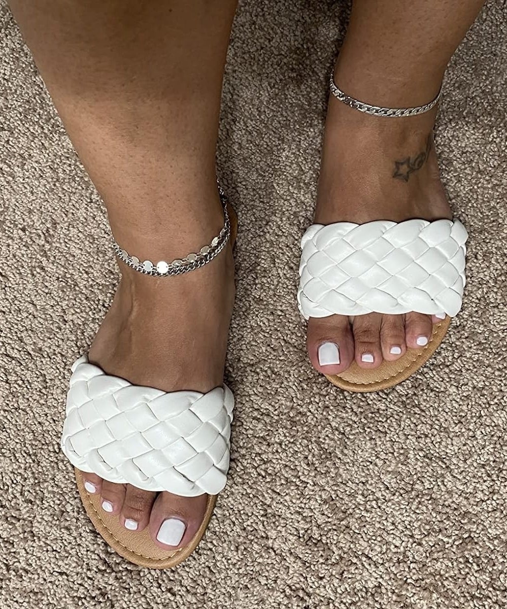 a reviewer&#x27;s feet in white braided slide sandals with an ankle bracelets, on a carpeted floor