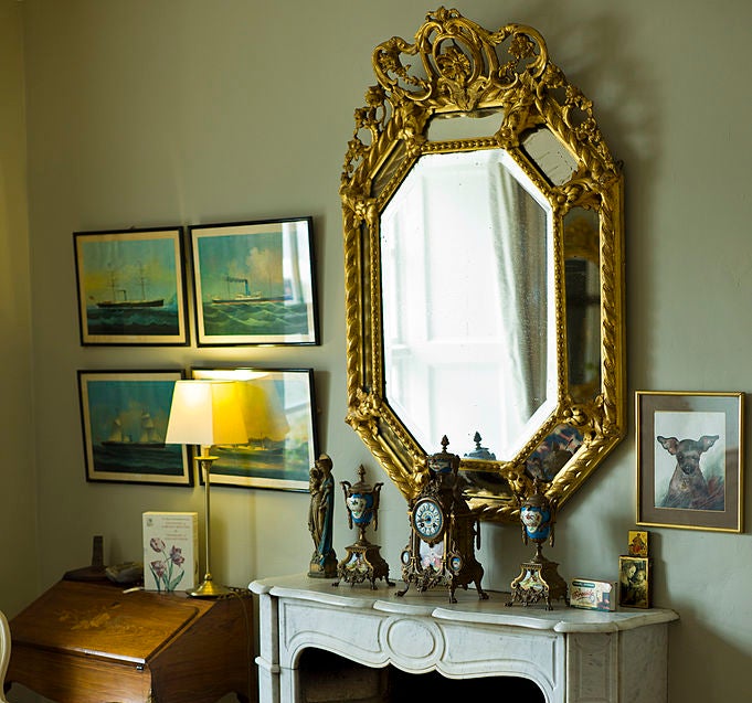 Elegant room with an ornate mirror above a fireplace, surrounded by artwork and a vintage writing desk