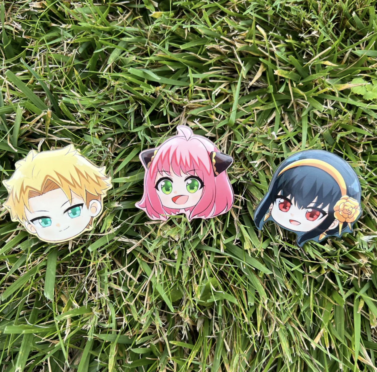 Three anime character keychains are laid out on grass, available for purchase