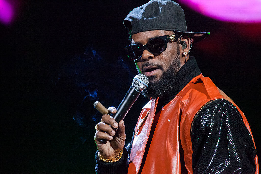 R. Kelly in a black hat and sparkly shirt holds a microphone and cigar onstage