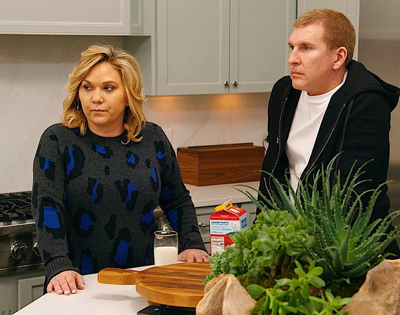 Julie and Todd Chrisley are seated at a kitchen counter with a thoughtful expression, surrounded by everyday kitchen items