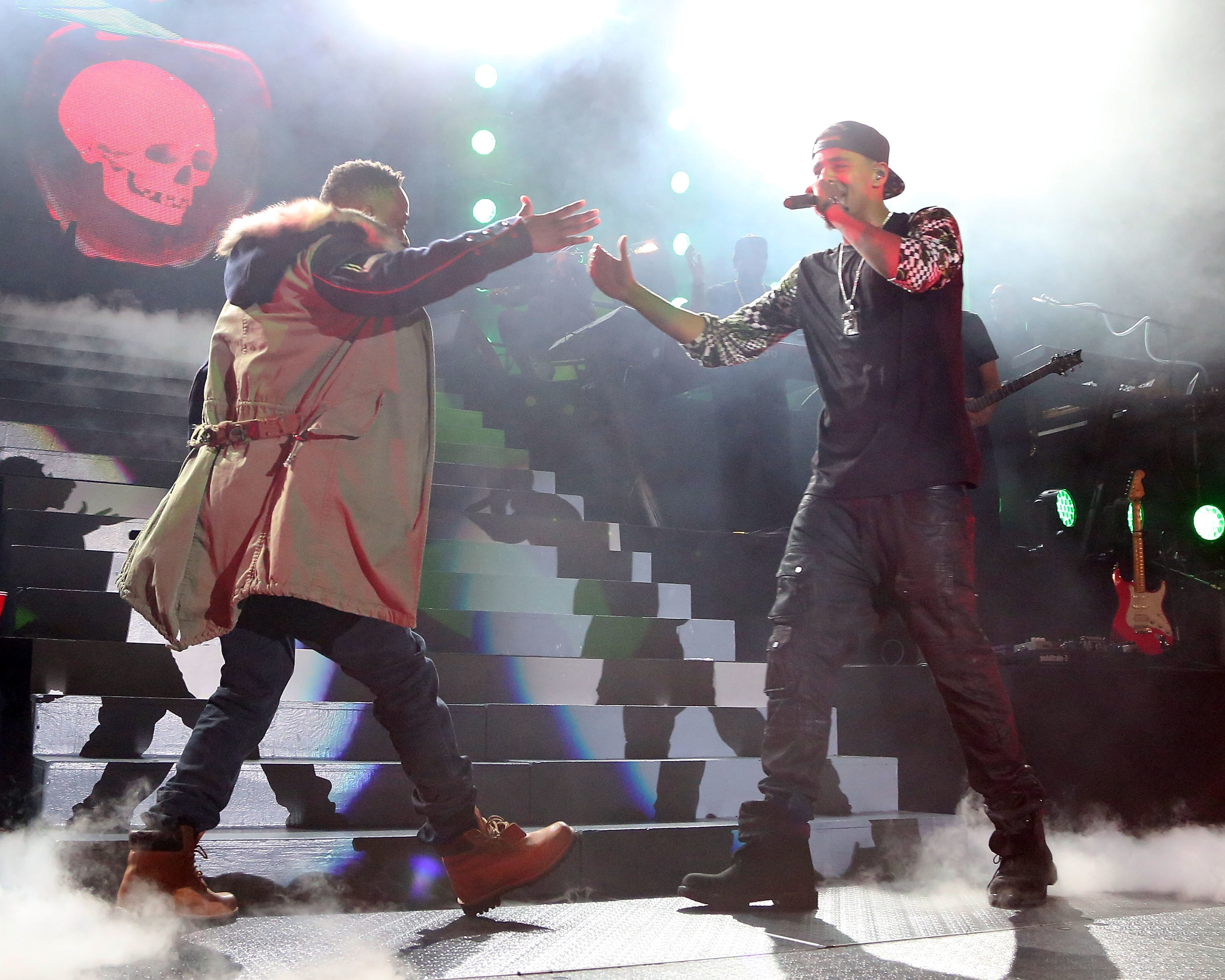 Two musicians on stage, one reaching out to the other, both in casual performance attire with graphic elements, amid stage fog and lights