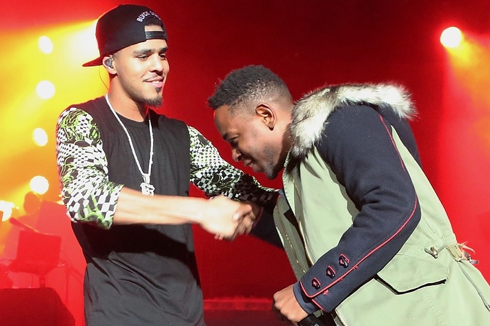Two artists on stage, one in a printed jacket and cap, the other in a coat with red detailing, shaking hands