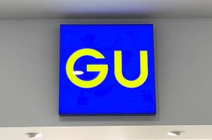 Sign with the letters "GU" on a blue background, mounted on a wall with ceiling lights visible