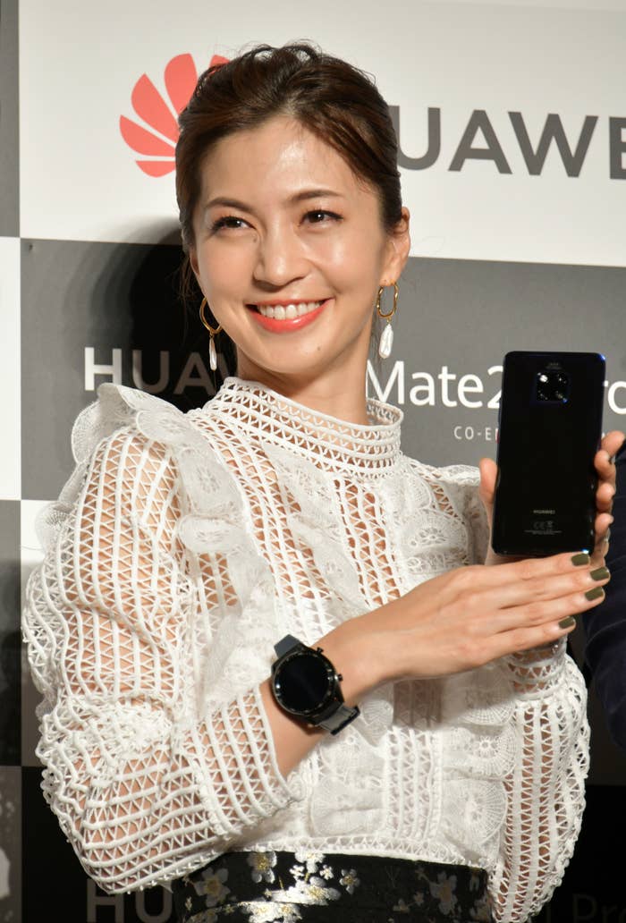 女性がホワイトレースのトップスを着てスマートフォンを持って笑顔でポーズを取っています。