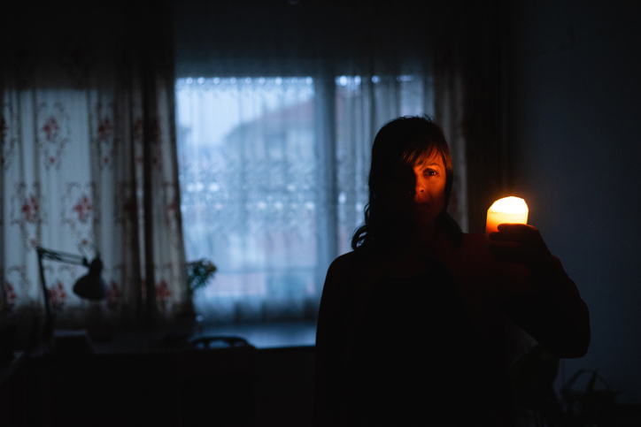 Woman in a dark room holding a lit candle near her face, with curtains and a window in the background