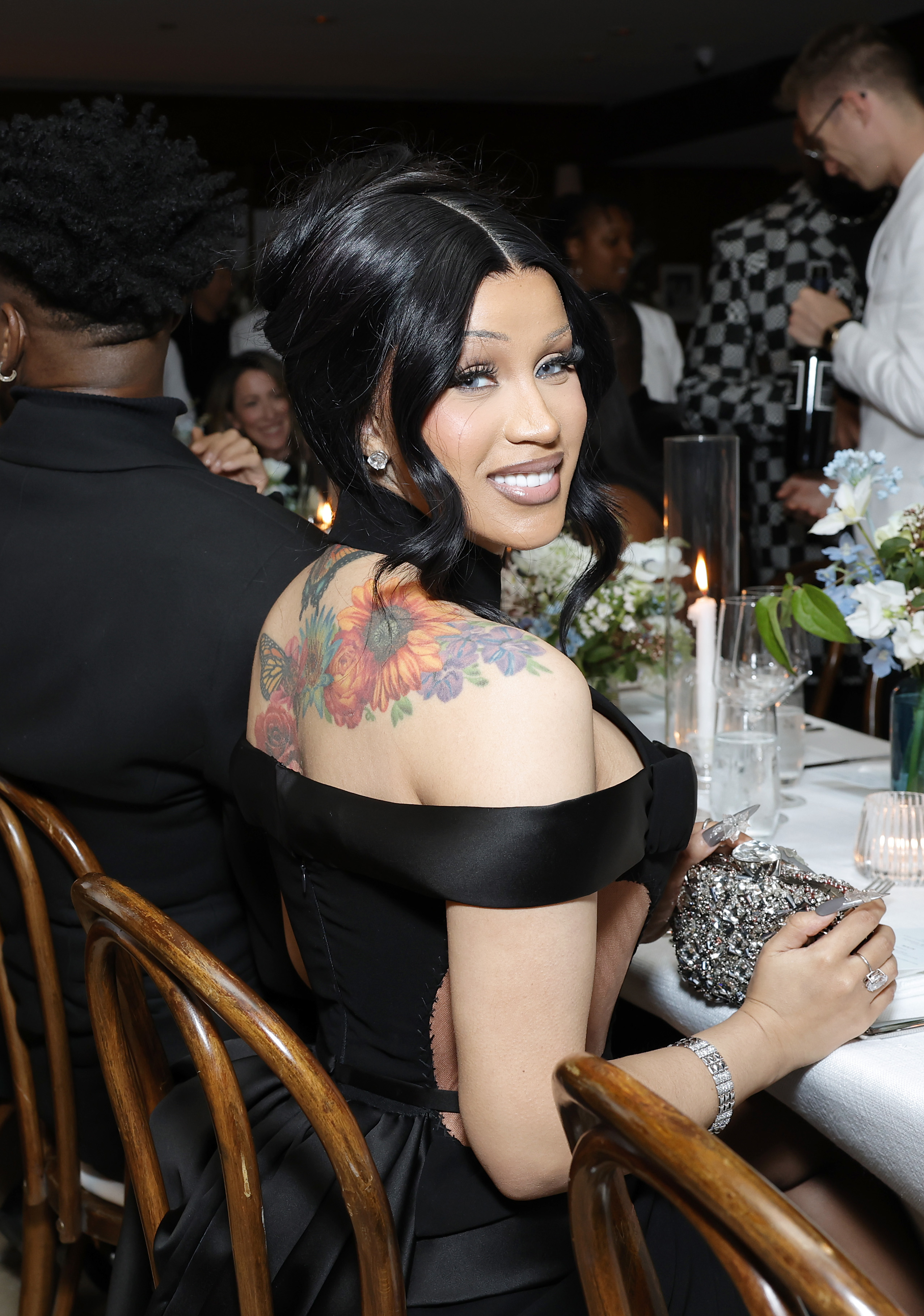 Cardi B in a black off-shoulder gown with floral tattoo visible, smiling at an event