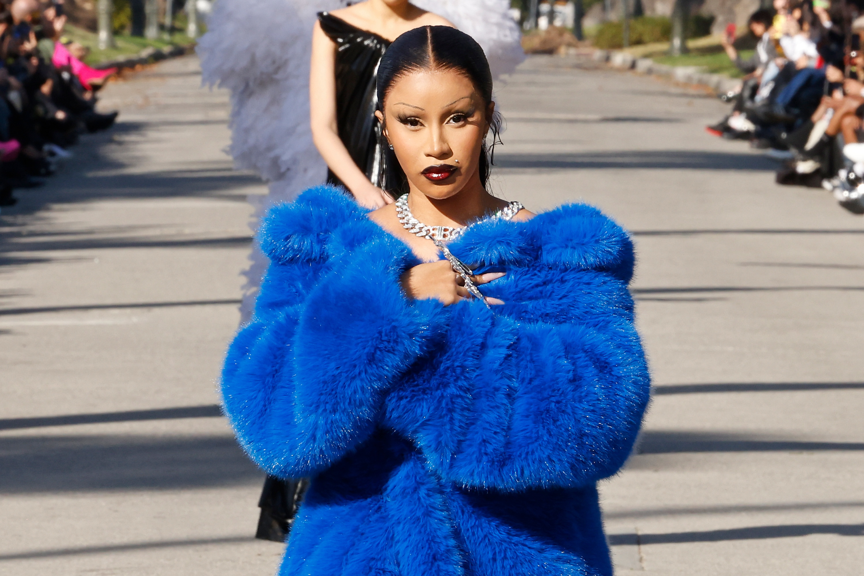Cardi B in an oversized blue fur coat, hands clasped near neck, posing outdoors