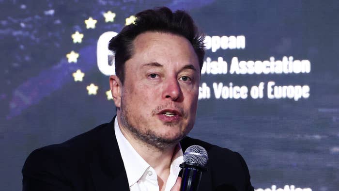 Elon Musk speaking into a microphone at an event with a starry background