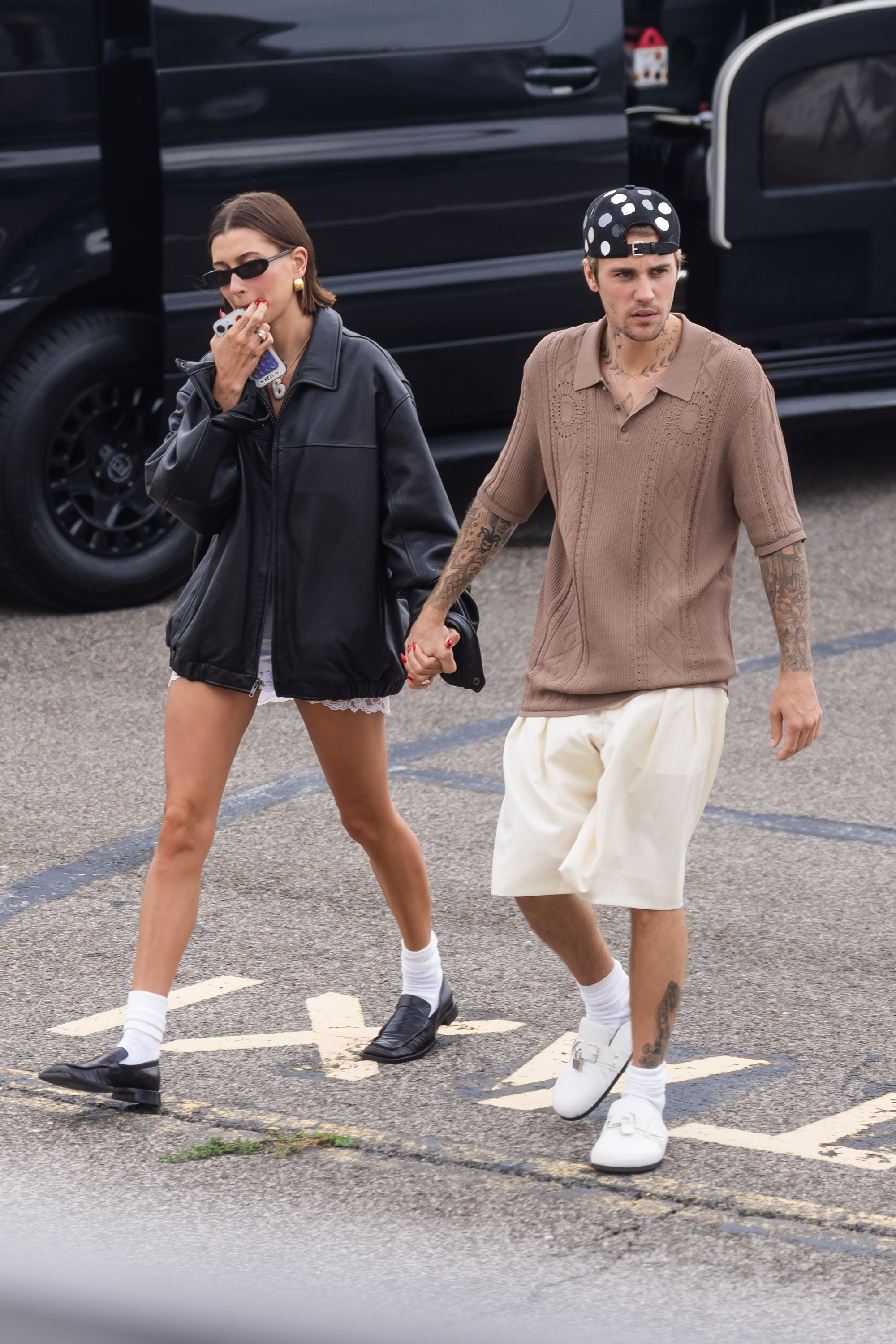 Hailey Bieber and Justin Bieber walking hand in hand, casual attire; Hailey with shorts and an oversized shirt, Justin in a cap and shorts