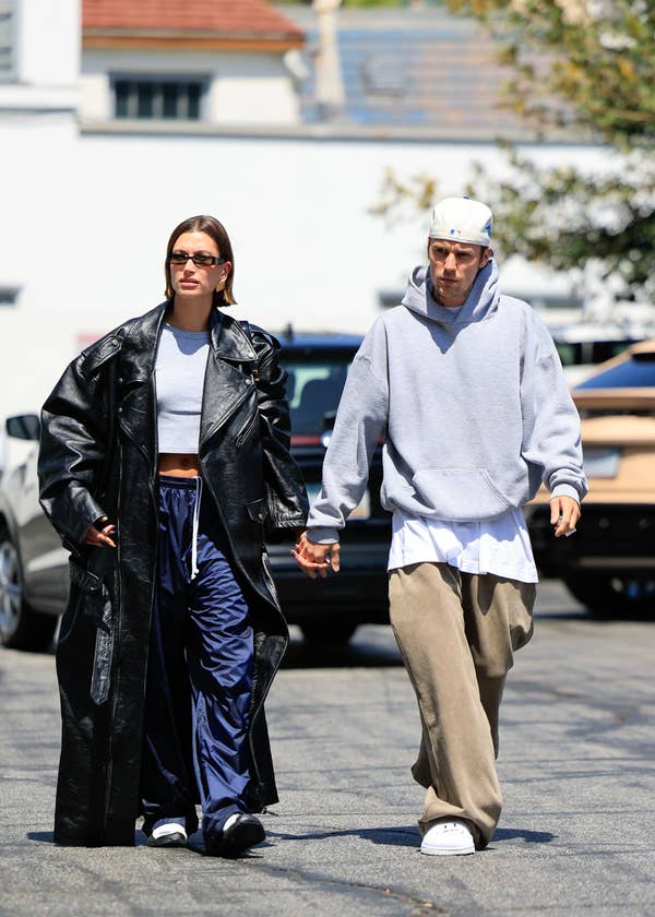 Two individuals walking side by side; one is in a long coat and sunglasses, the other in a hoodie and cap